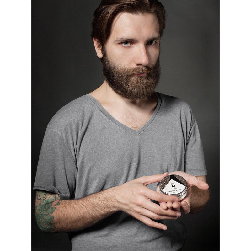 Man with a beard, tattoo, and dark hair, holding a tin of Beard Reverence beard balm. 